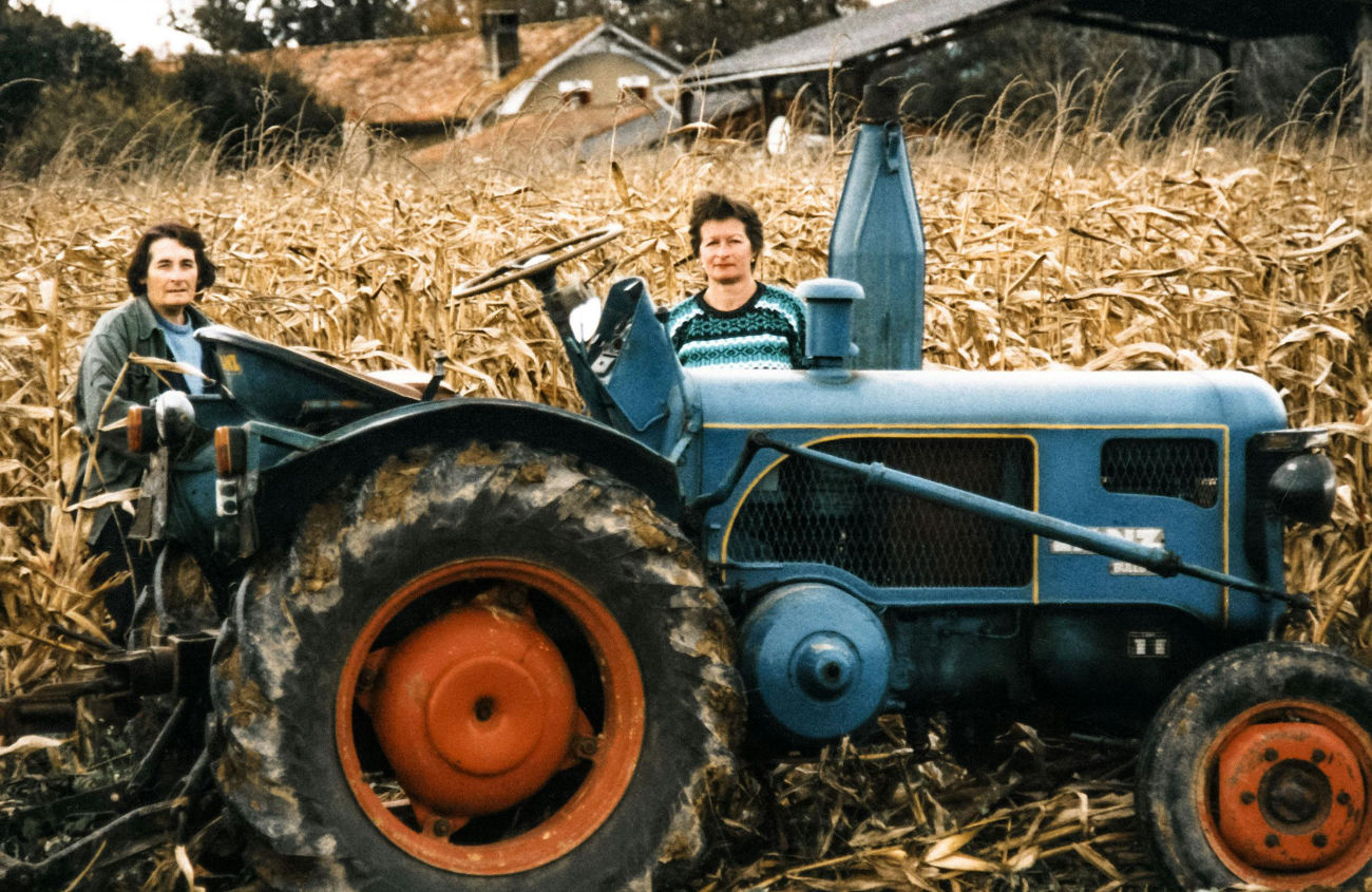Duas senhoras no campo, no processo de cultivo e recolha, junto a um trator. Imagem que representa o trabalho da sementeira à colheita.